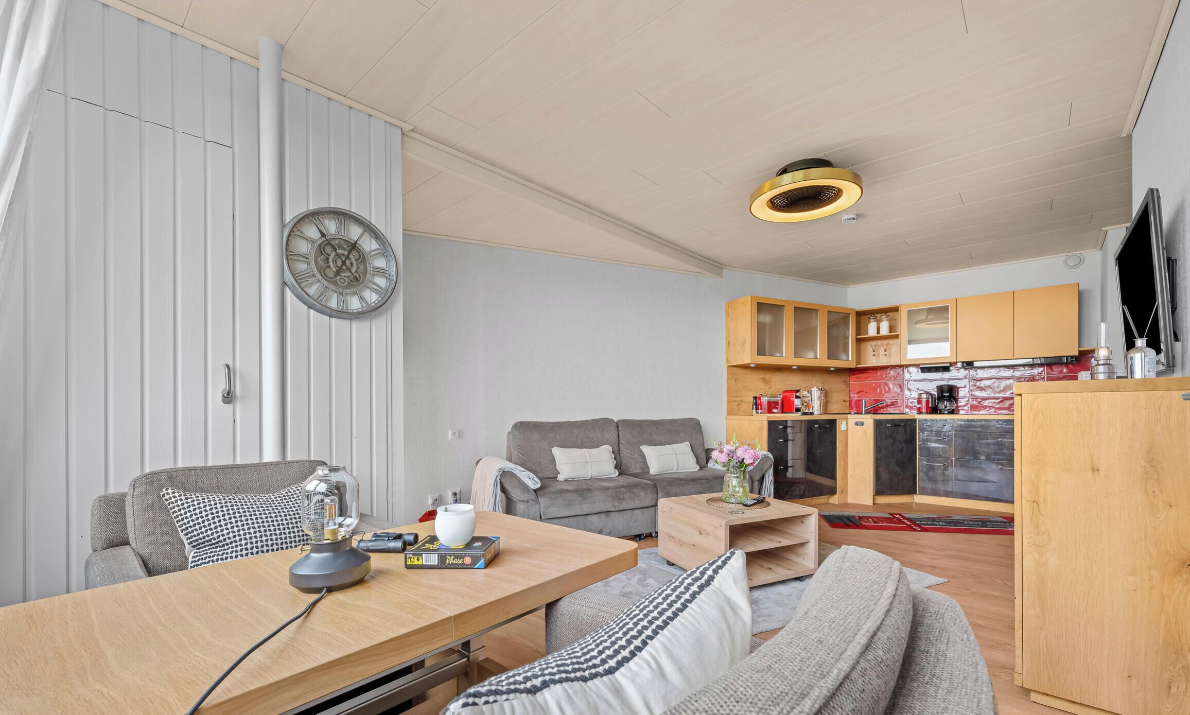 A modern living room with a sofa, a dining table, a kitchen area with cabinets, and a wall clock.