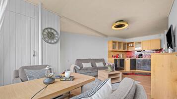A modern living room with a sofa, a dining table, a kitchen area with cabinets, and a wall clock.