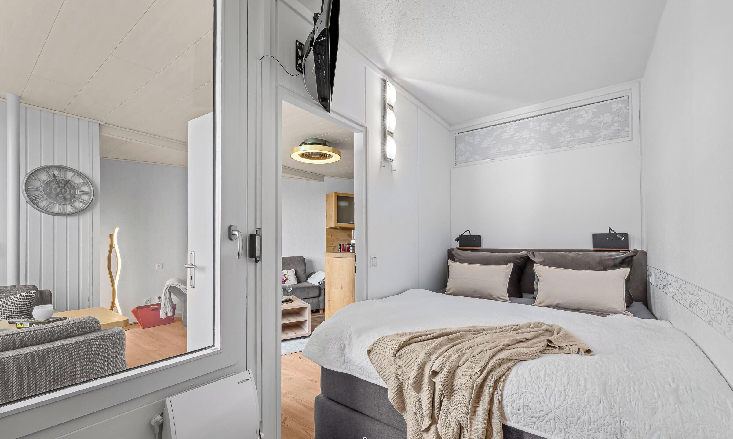 A modern bedroom with a bed, bedside tables, a clock, and a view of the adjacent living area.