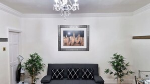 Una sala de estar con un sofá negro, dos plantas en macetas, una araña y una imagen enmarcada de un paisaje urbano en la pared.