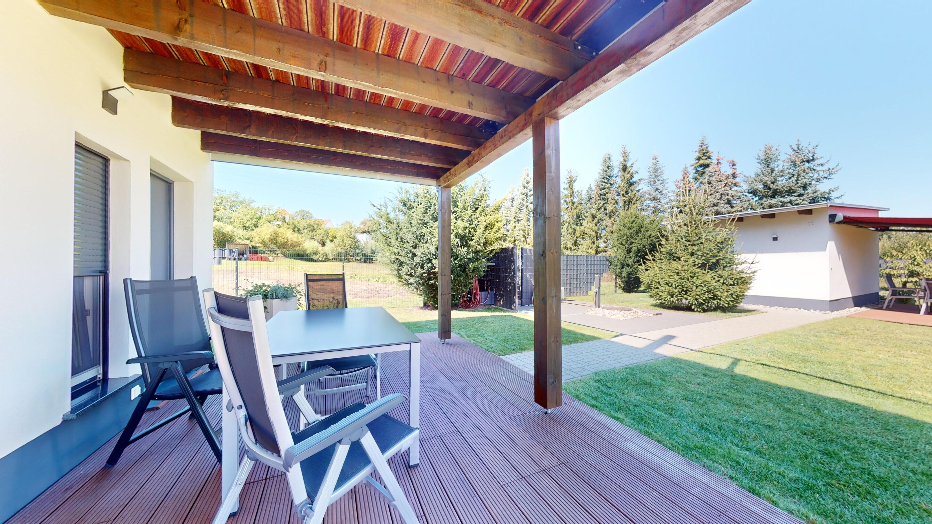 An outdoor patio with a table and chairs, a wooden pergola, and a view of a backyard with a lawn and trees.