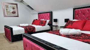Habitación de hotel con tres camas, cada una con cabecera roja, ropa de cama blanca y almohadas con estampado rojo y negro. Hay una obra de arte enmarcada en la pared y una mesita con una lámpara.