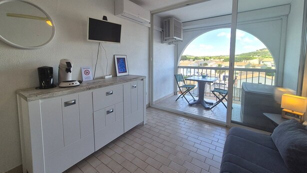 Ein Balkon mit Tisch und Stühlen, einer Couch, einem an der Wand befestigten Fernseher und einem Schrank mit einer Kaffeemaschine.