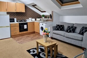 Eine kompakte Küche mit Mikrowelle, Herd und Waschmaschine. Ein graues Sofa mit gemusterten Kissen und ein kleiner Holztisch mit Blumenvase.
