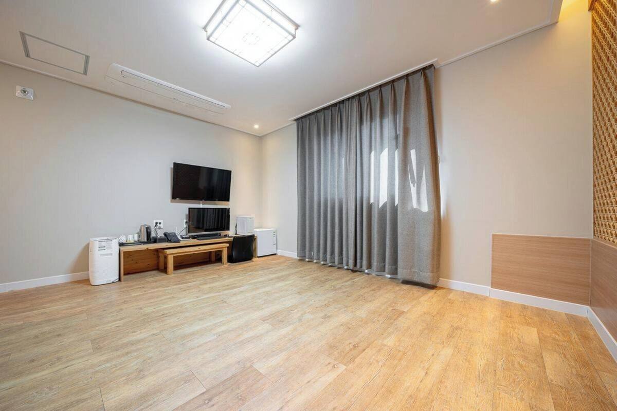 A room with a wooden floor, a flat-screen TV mounted on the wall, a desk with a chair, a small heater, and a large window with curtains.