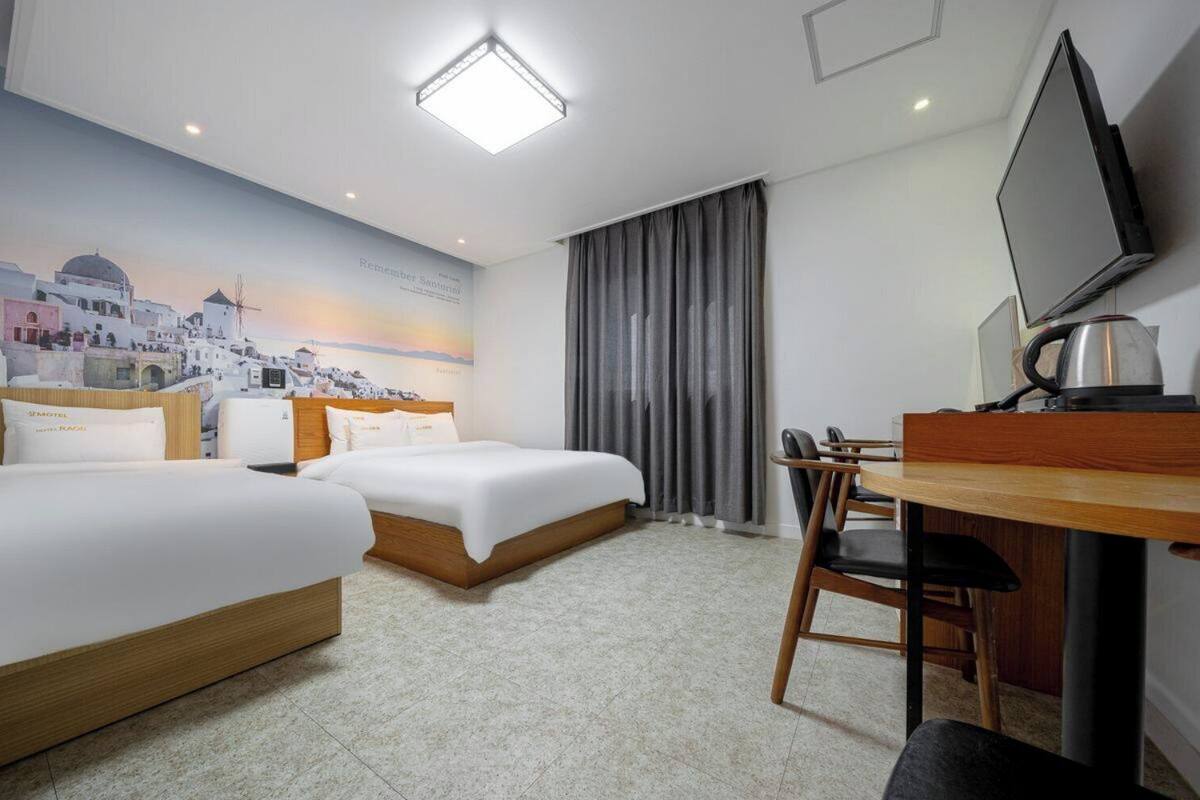 A hotel room with two beds, a desk, a chair, a television, and a mural of a coastal town.