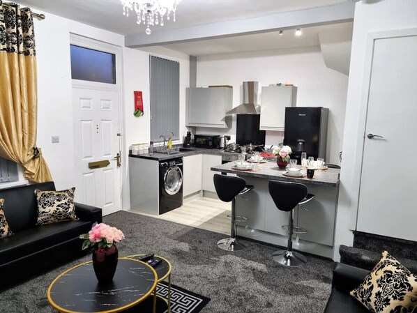 A modern kitchen with a central island, a dining area with two bar stools, a sofa set, and a washing machine.