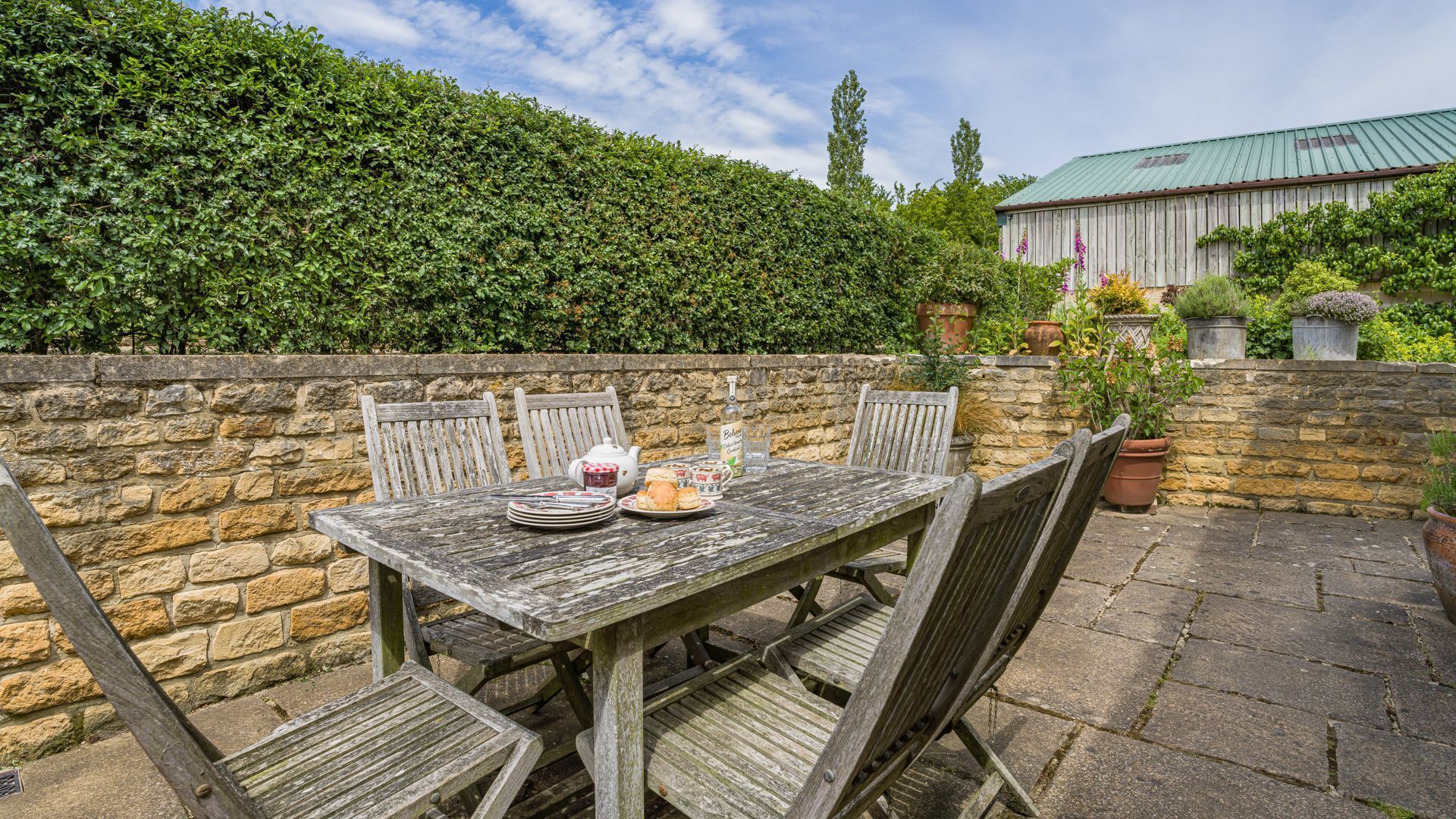 A rustic outdoor dining area with a stone wall, wooden table, and chairs.