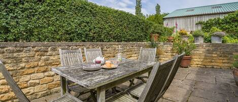 A rustic outdoor dining area with a stone wall, wooden table, and chairs.
