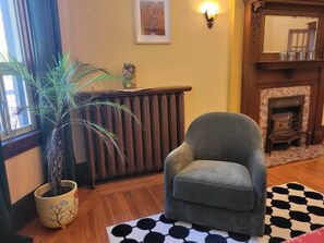 A room with a grey armchair, a potted plant, a fireplace, and a patterned rug. - Victorian on Herkimer (Hamilton)