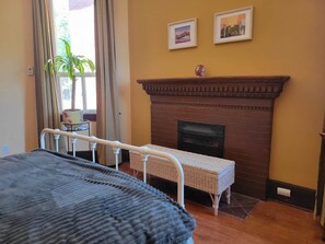 A bedroom with a bed, a fireplace, a window with curtains, and two framed pictures on the wall.