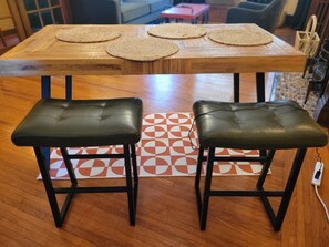 A wooden dining table with three woven placemats and two black leather stools.