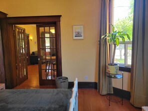 A room with a bed, a window with curtains, a plant on a side table, and a door leading to another room.