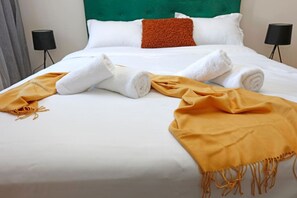 Ein ordentlich bezogenes Bett mit weißen Bettwäsche, einer senffarbenen Tagesdecke und aufgerollten Handtüchern.