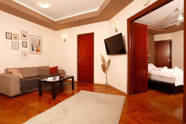Un salon comprenant un canapé, une table basse, un téléviseur fixé au mur et une chambre à coucher adjacente.