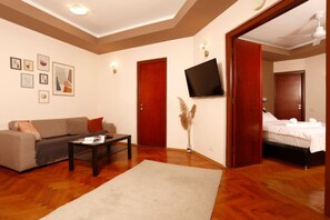 Ein Wohnzimmer mit einer Couch, einem Couchtisch und einem an der Wand montierten Fernseher. Zwei Türen führen zu einem Schlafzimmer mit einem Bett.