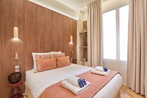 Ein Hotelzimmer mit einem großen Bett, einem Nachttisch mit einer Schale, einer Wandlampe, einem Fenster mit Vorhängen und einem Regal mit einer Pflanze.