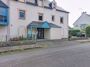 A two-story building with a blue entrance canopy, a stone wall with flowers, and a car parked on the street. - Claudel by Interhome (Saint Malo)
