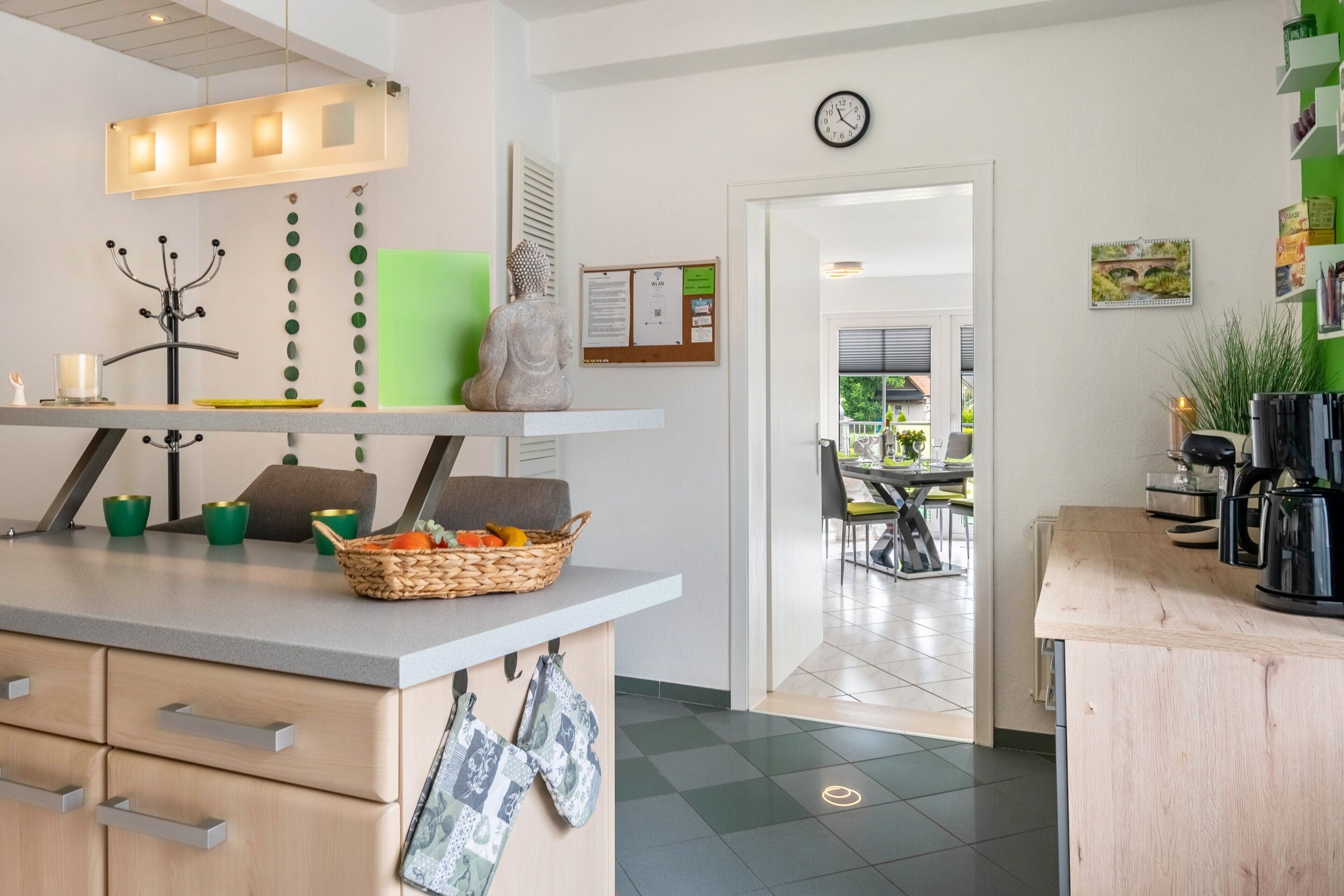 A modern kitchen with a light wood countertop, a green cutting board, a basket of fruit, and a wall-mounted clock.