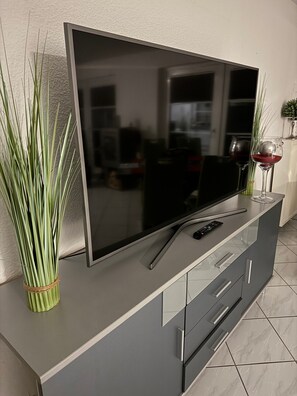 A flat-screen TV on a grey cabinet with a remote control, a glass vase with green plants, and a wine glass.