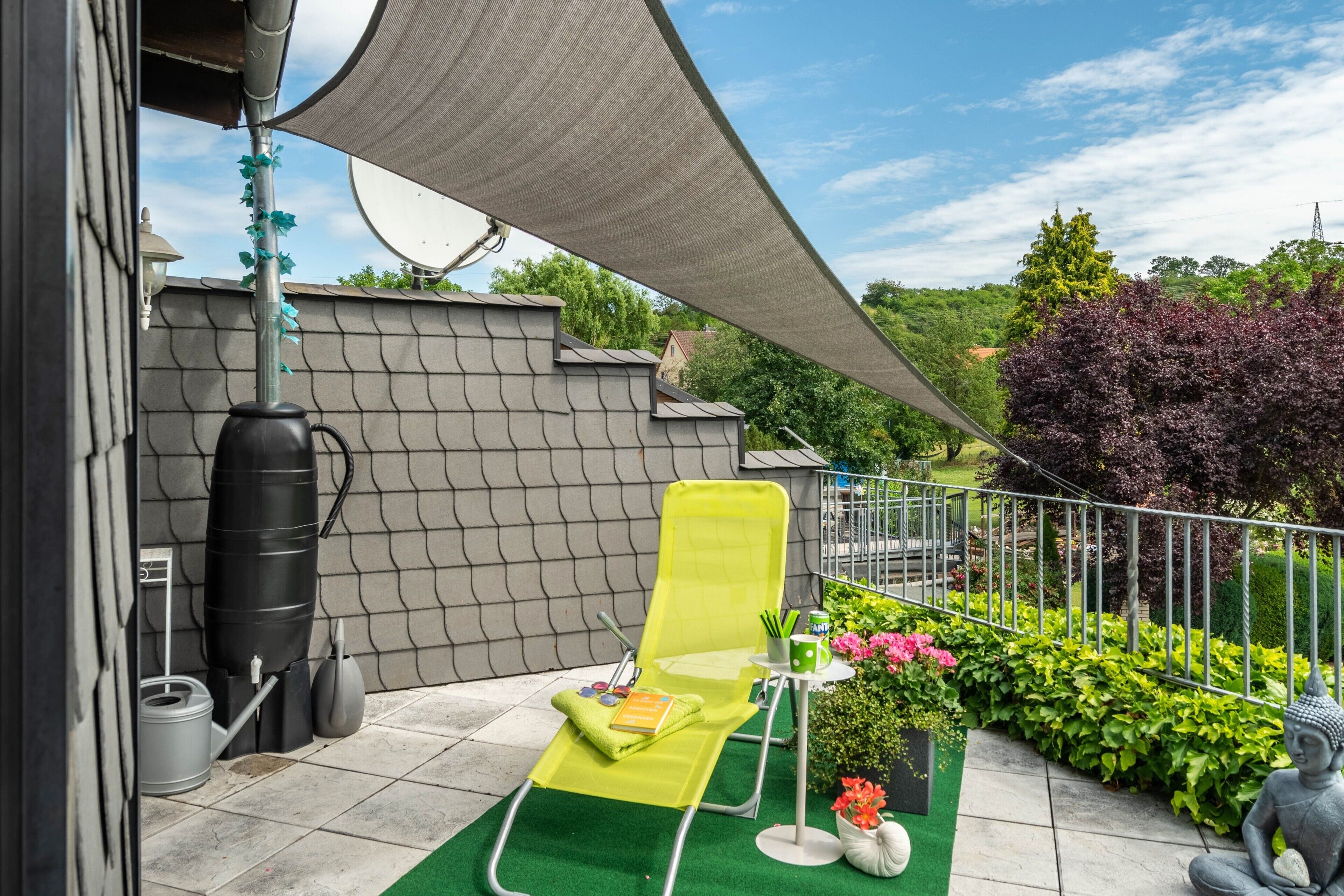 Un balcon avec une chaise longue verte, une petite table sur laquelle se trouve une tasse verte et une statue.