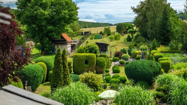 Un jardín cuidado con diversas formas de seto, un pequeño edificio y un banco.