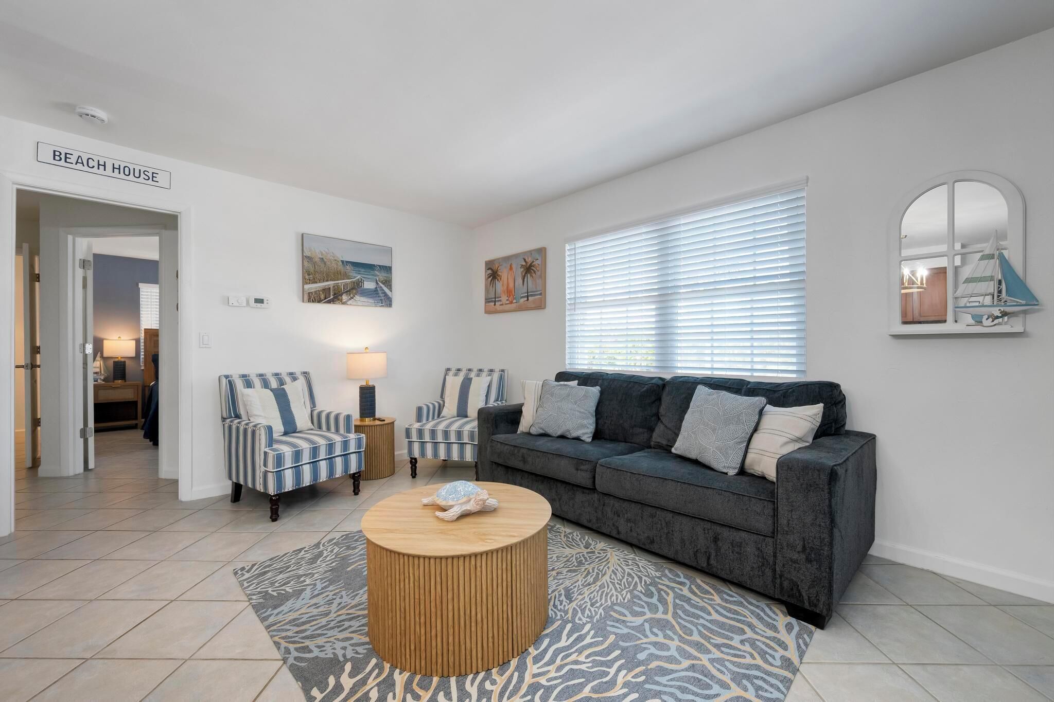 Ein Wohnzimmer mit einer Couch, zwei Sesseln, einem Couchtisch und einem Teppich. Eine Tür mit der Aufschrift 'BEACH HOUSE' und ein Fenster mit Jalousien sind zu sehen.