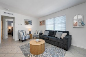 Ein Wohnzimmer mit einer Couch, zwei Sesseln, einem Couchtisch und einem Teppich. Eine Tür mit der Aufschrift 'BEACH HOUSE' und ein Fenster mit Jalousien sind zu sehen.