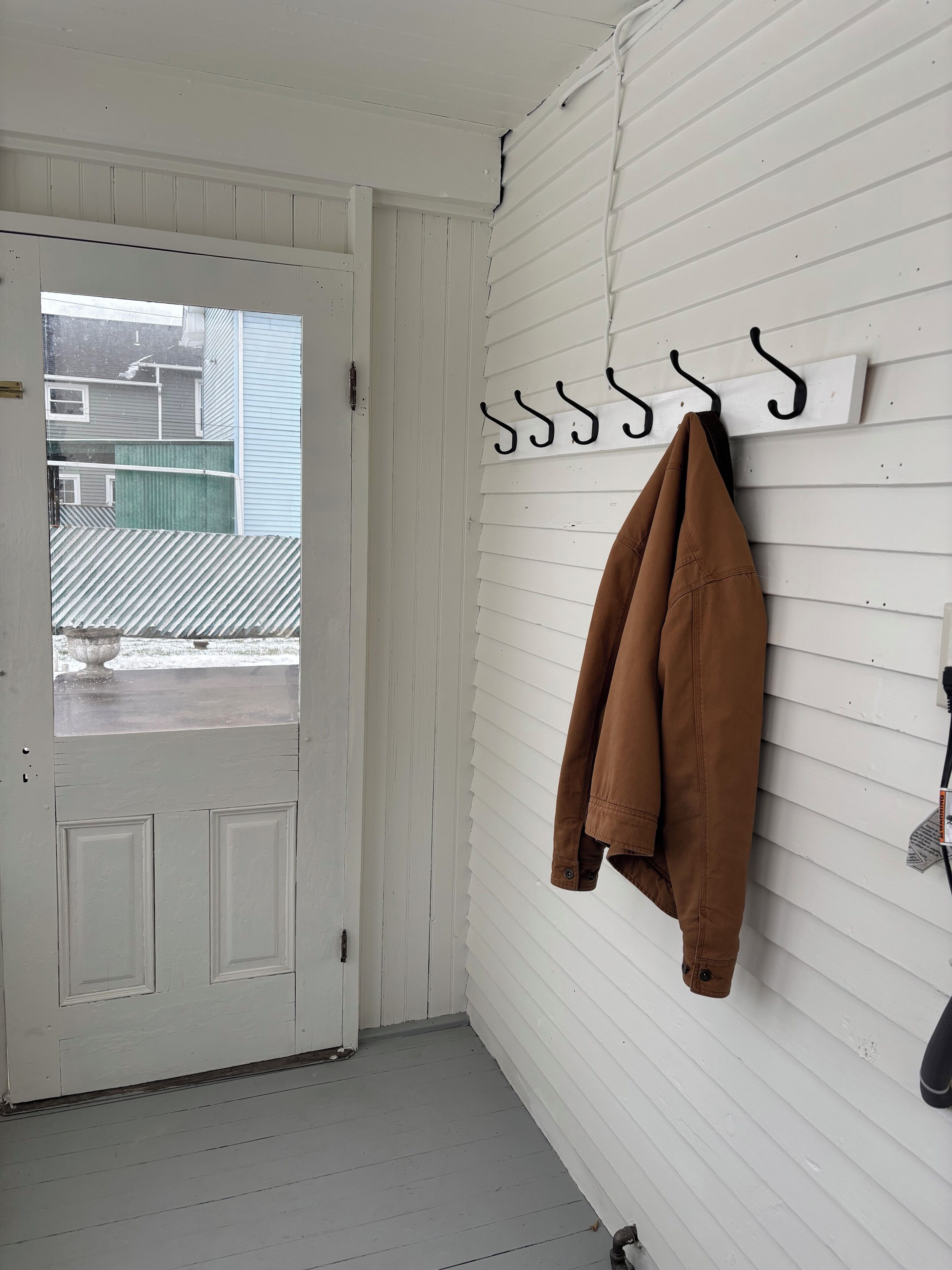 A white door with a glass panel, a brown jacket hanging on a wall-mounted hook, and a white wall with horizontal siding.