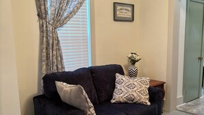 Un sofá oscuro con cojines estampados, un jarrón decorativo con flores y un cartel enmarcado en la pared.