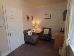 A room with two armchairs, a small table with a lamp, a wooden side table, a framed picture on the wall, a door, and a window with curtains. - Single room-Economy-Ensuite-Mountain view (Crianlarich)