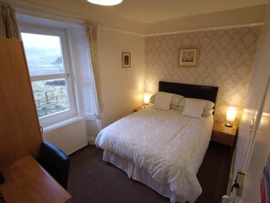 A bedroom with a bed, two bedside lamps, a window with curtains, a radiator, and a framed picture on the wall. - Single room-Economy-Ensuite-Mountain view (Crianlarich)