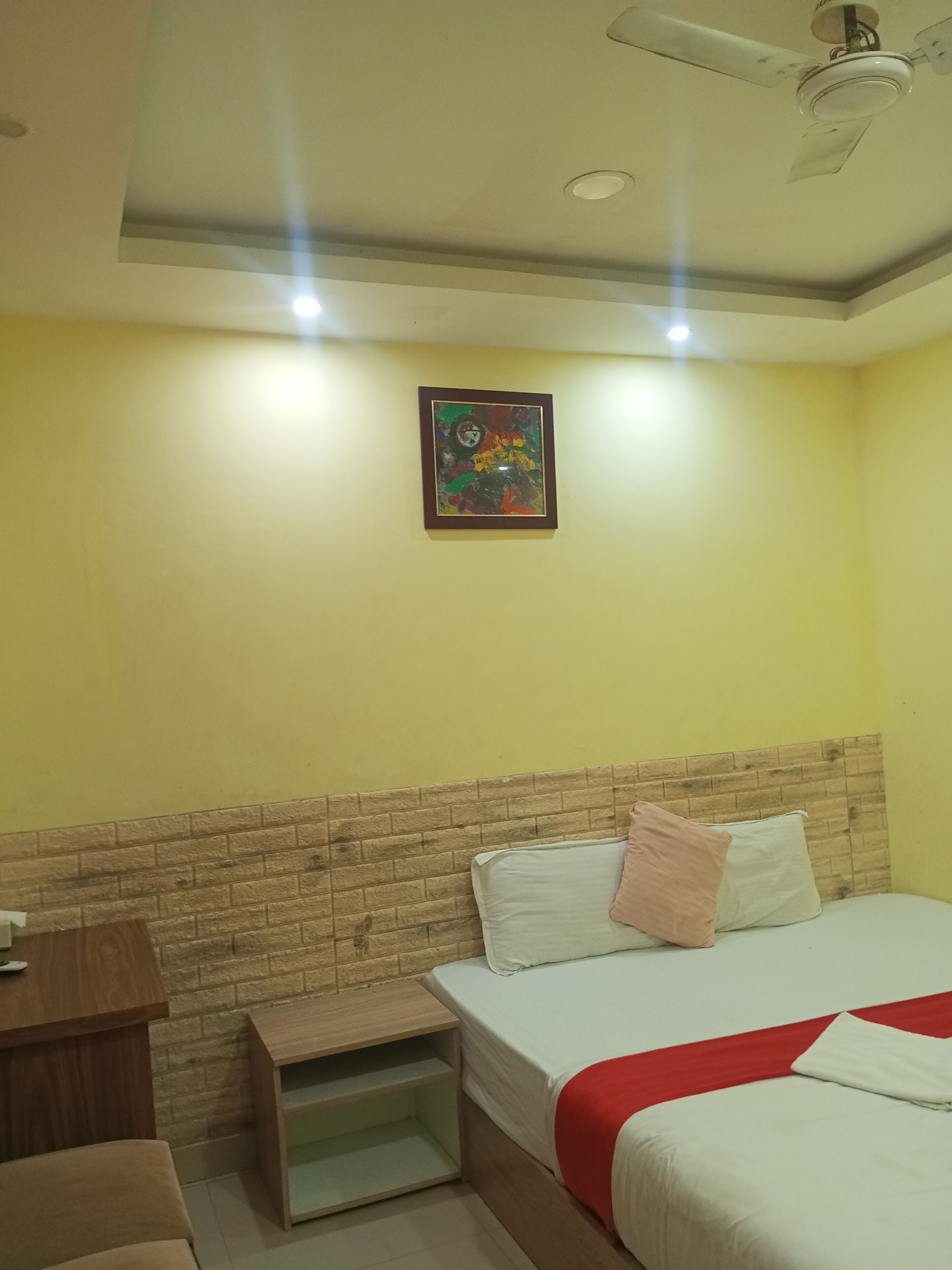A hotel room with a brick-patterned headboard, a bed with white and red bedding, a small wooden nightstand, a ceiling fan, and a colorful abstract painting on the wall.