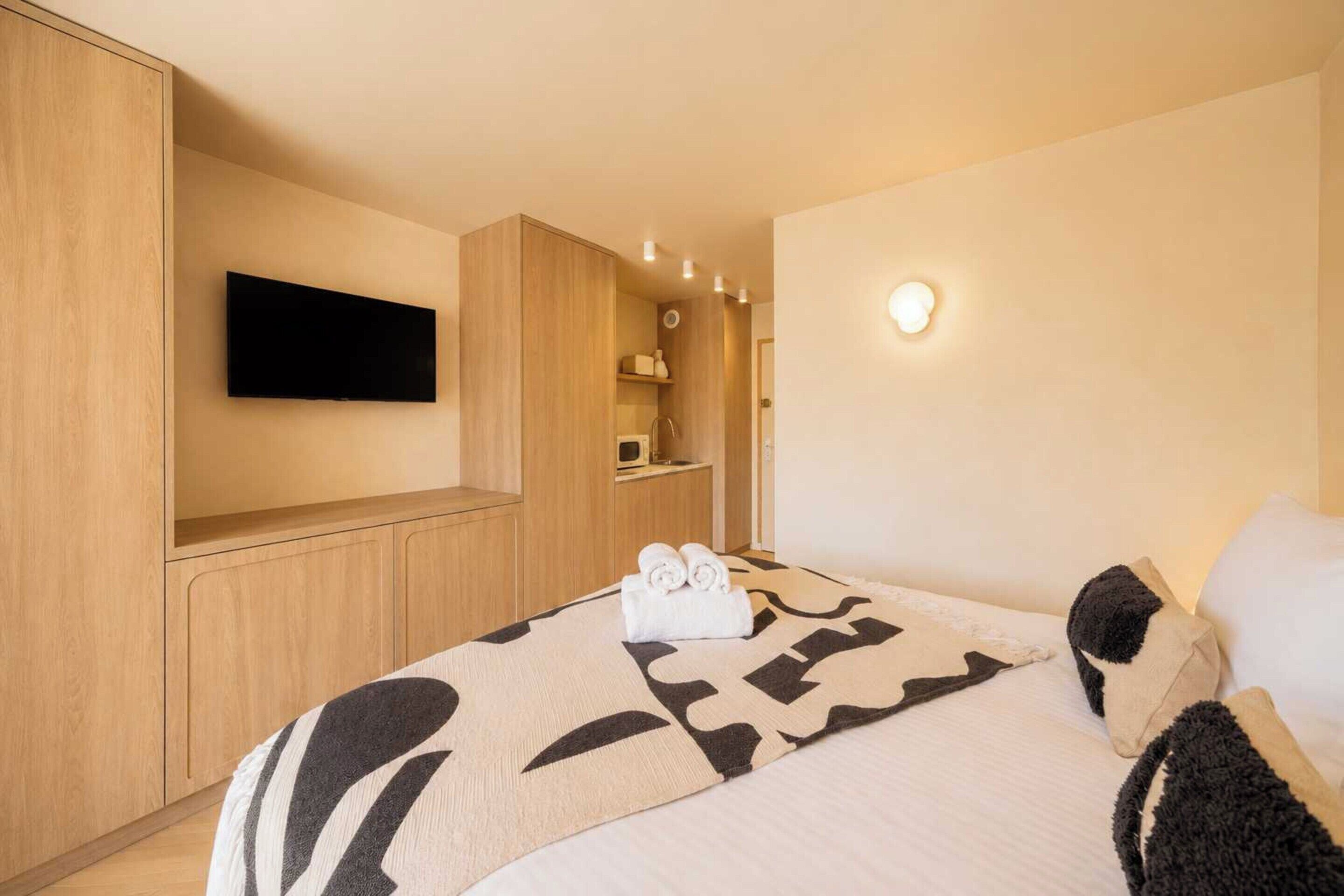 Una habitación de hotel moderna con televisión de pantalla plana, una cama con manta estampada, un estante pequeño con artículos de aseo y una lámpara fijada a la pared.