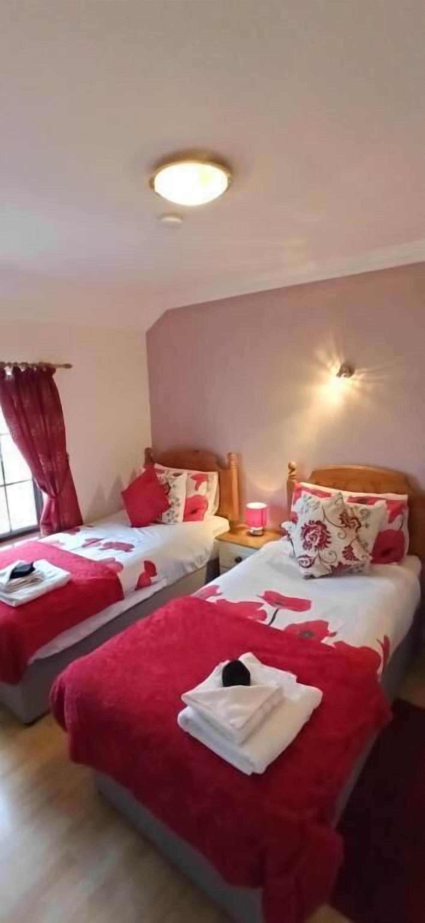 Habitación de hotel con dos camas, cada una con ropa de cama roja y almohadas estampadas, una mesita de noche con lámpara, y una ventana con cortinas rojas.