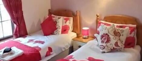 Habitación de hotel con dos camas, cada una con ropa de cama roja y almohadas estampadas, una mesita de noche con lámpara, y una ventana con cortinas rojas.