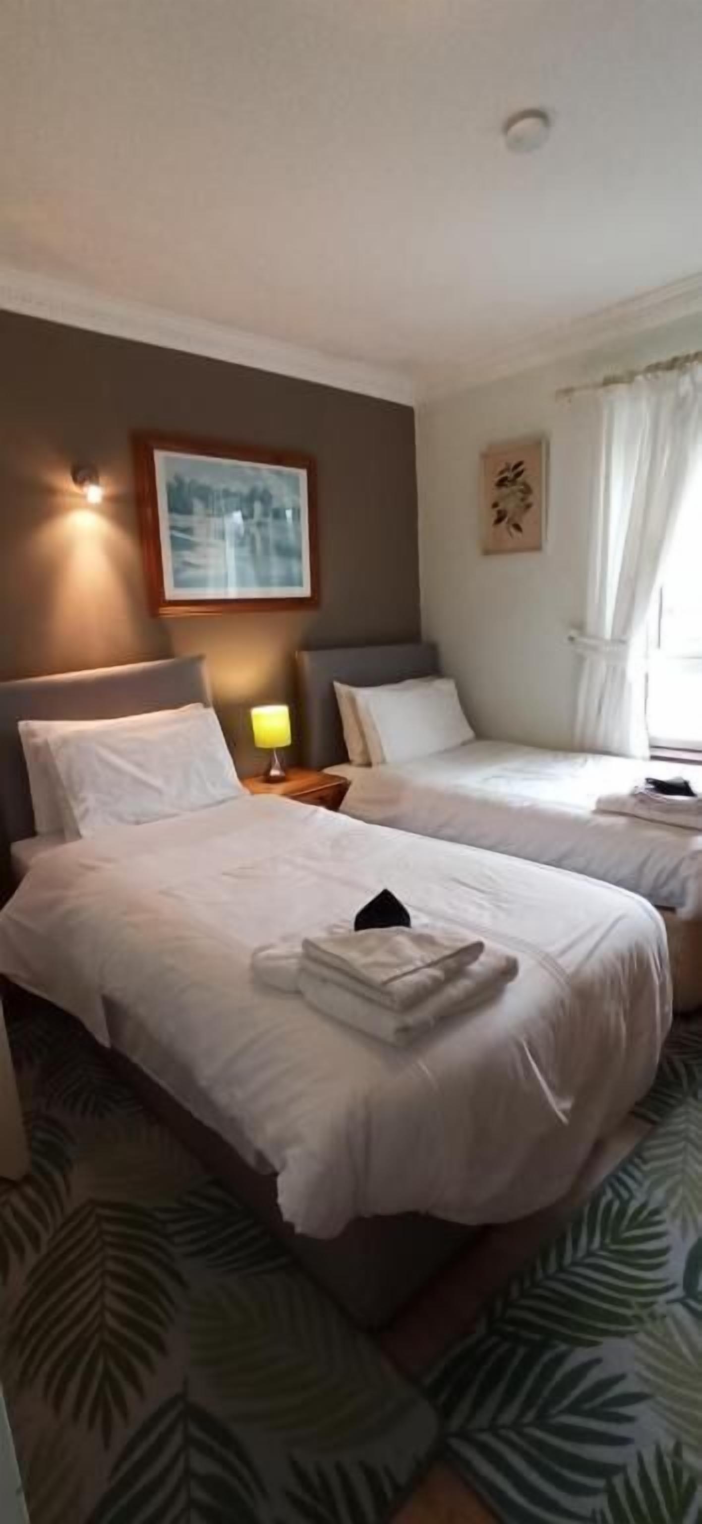 Quarto de hotel com duas camas, uma mesa de cabeceira com abajur, um quadro na parede e uma janela com cortinas.