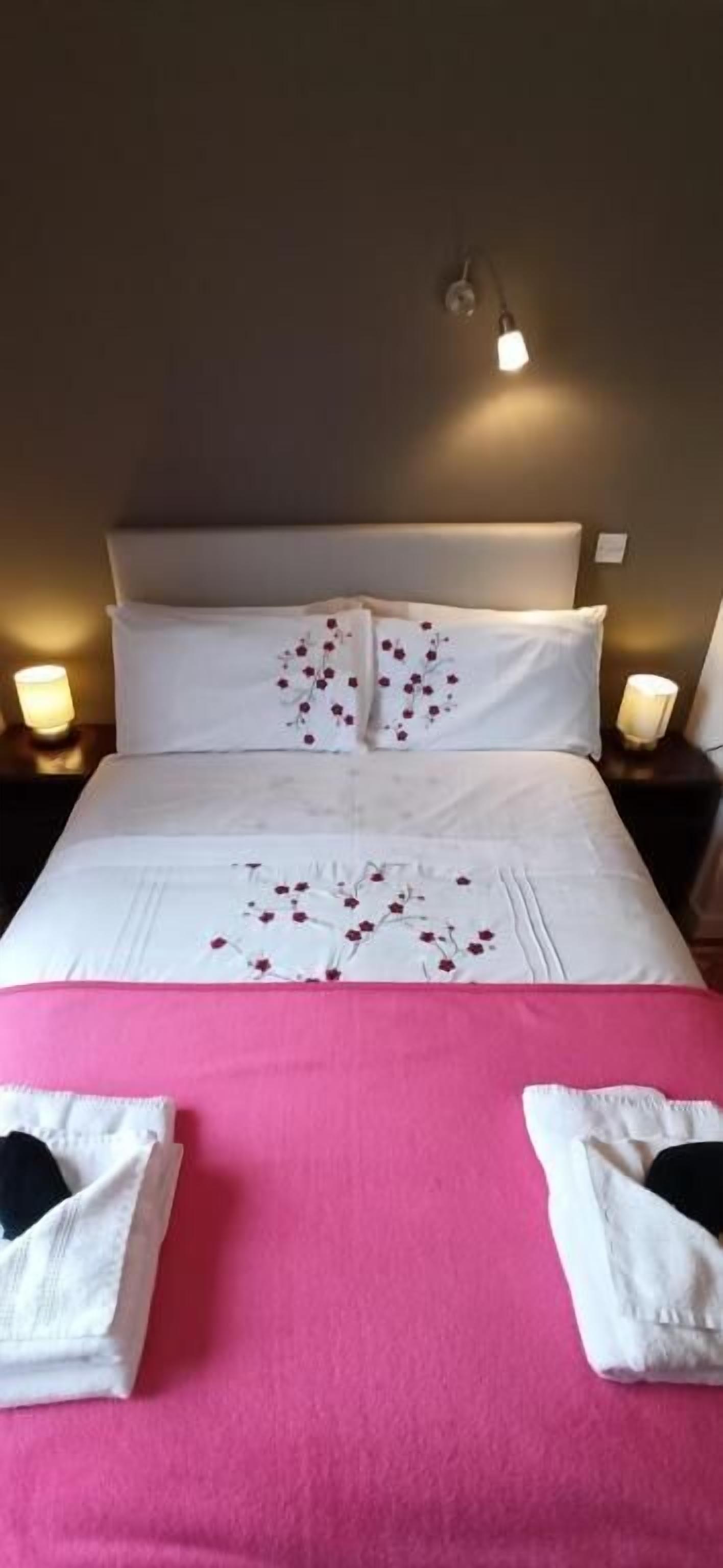 Cama bem arrumada com lençóis brancos e colcha rosa, duas toalhas brancas sobre a cama e luminárias de cabeceira de cada lado.