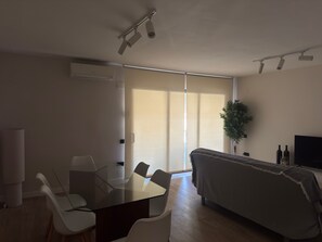 Una sala de estar moderna con una mesa de comedor de cristal, sillas blancas, un sofá y un ventanal grande.