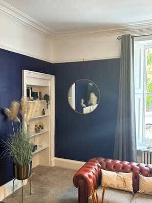 A room with a dark blue wall, a round mirror, a red leather sofa, and a bookshelf with various items. - Greyfriars Garden No (14A) | St Andrews (St Andrews)