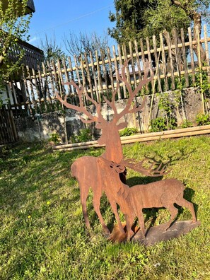 Escultura de cerví metal·licà en una zona enjardinada amb una tanca de fusta i arbres al fons.