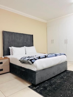 A bedroom with a bed, nightstand, wardrobe, and a black rug. - Room-airport Continental Guest House (Kempton Park)