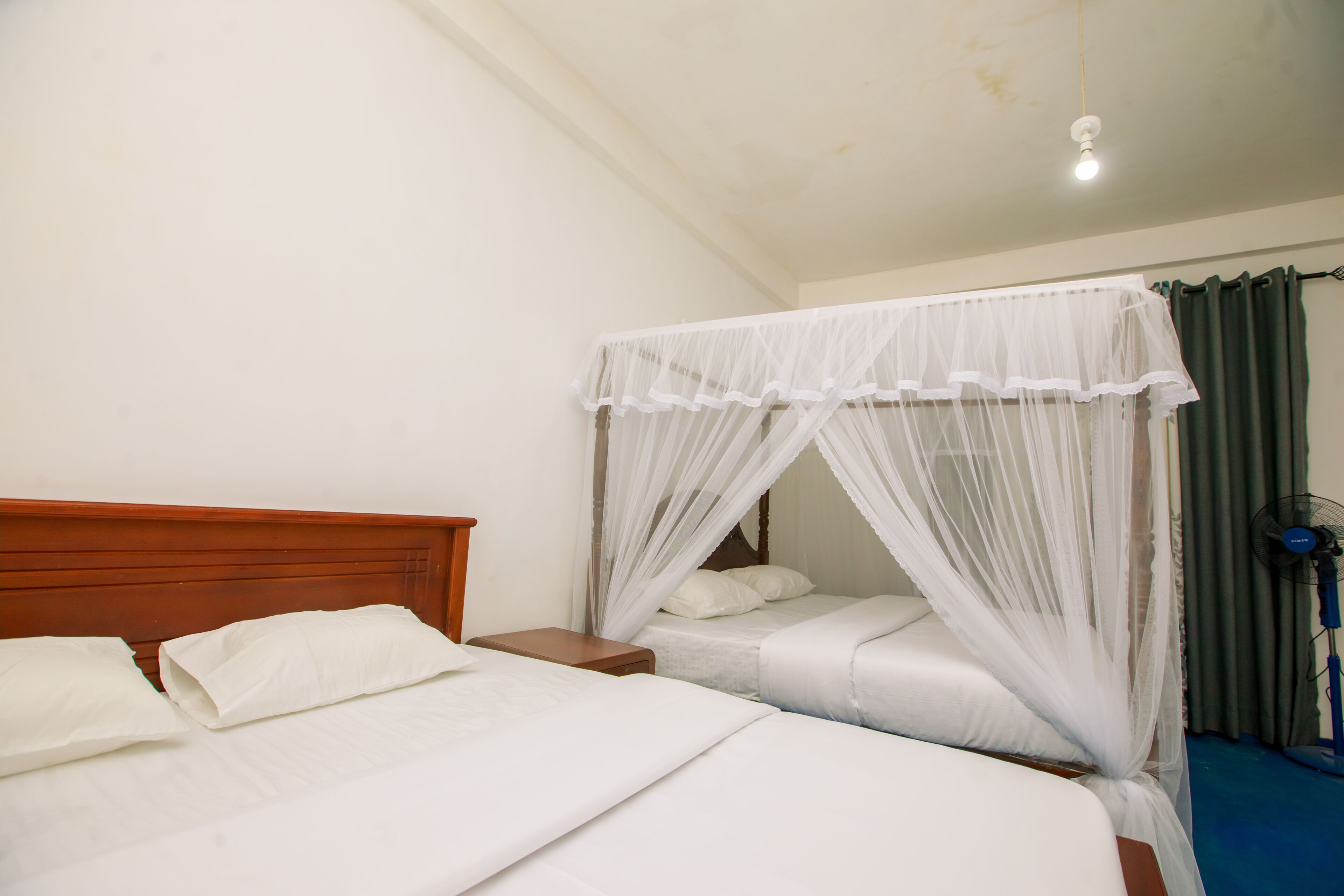 A bedroom with two beds, a wooden headboard, a bedside table, a ceiling fan, and a mosquito net over one bed.