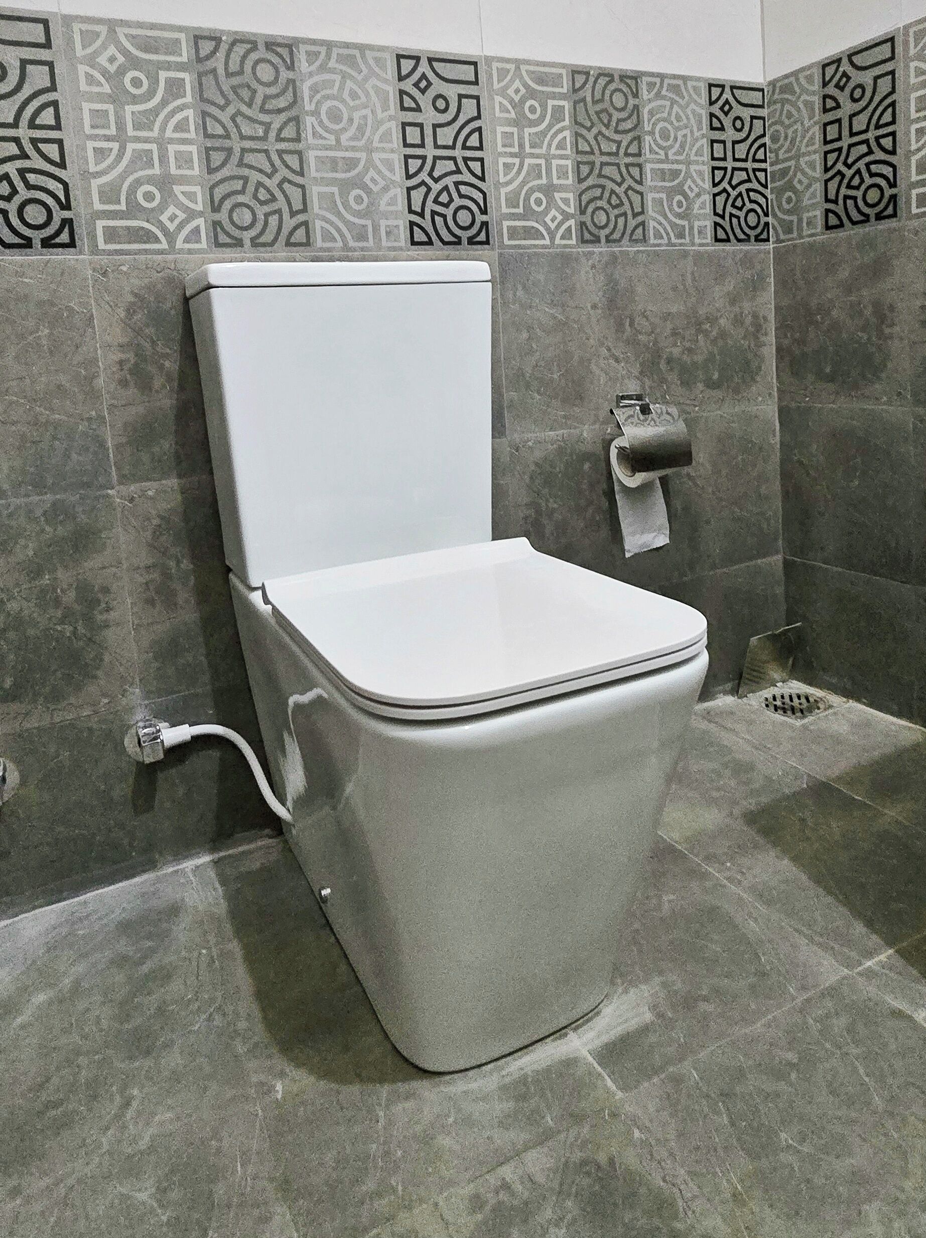 Des toilettes avec un abattant et un couvercle blancs, des carreaux gris sur les murs et un porte-rouleau de papier toilette fixé au mur.