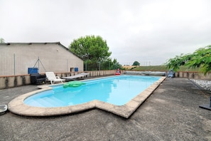 A swimming pool with a curved edge, surrounded by a concrete deck, a fence, and a building with a sloped roof. - Chambre Perle – 12 m² (Monclar-de-Quercy)