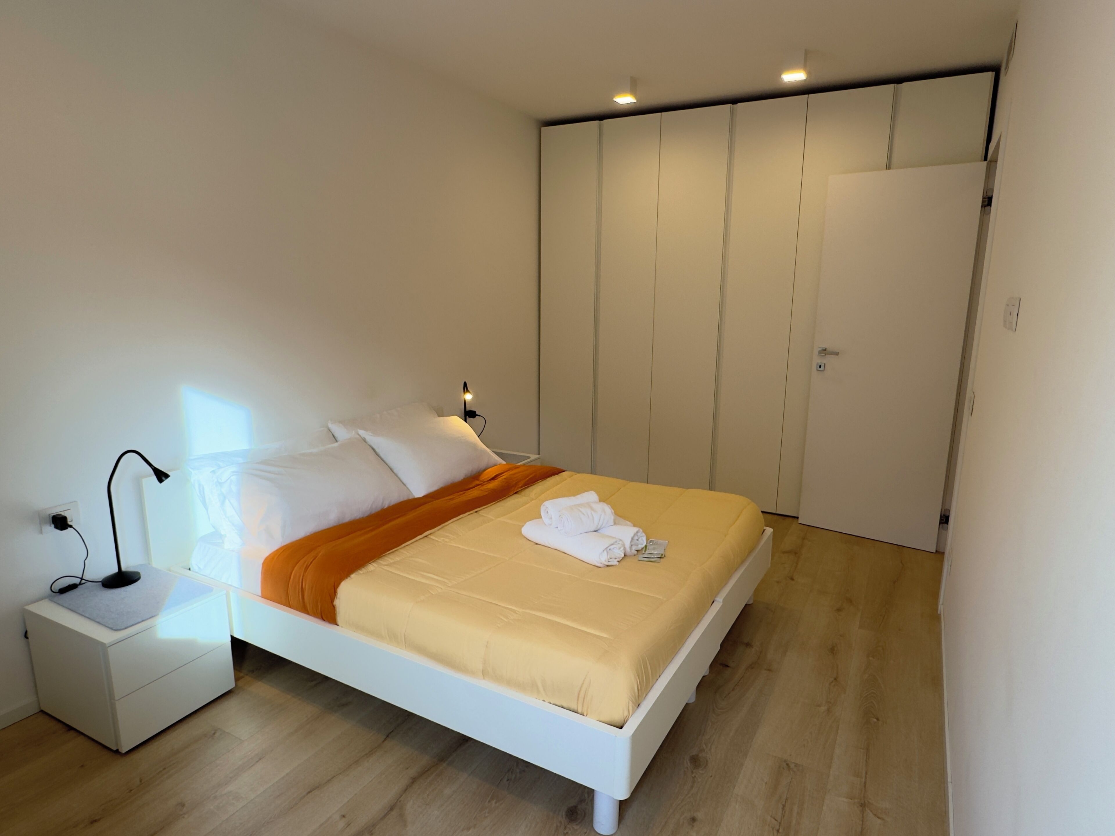 Un letto rifatto con un comodo poggiatesta bianco, un comodino con una lampada e un armadio con ante scorrevoli.