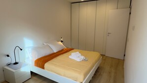 Een net opgemaakt bed met een wit hoofdeinde, een nachtkastje met een lamp en een inloopkast met schuifdeuren.