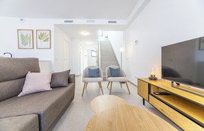 Ein modernes Wohnzimmer mit einer Couch, zwei Sesseln, einem Couchtisch aus Holz, einem Fernseher und einer Treppe, die zu einer oberen Etage führt.