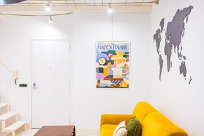 Een kamer met een witte deur, een gele bank, een wereldkaart aan de muur en een poster van het magazine 'The Napolitaner'.