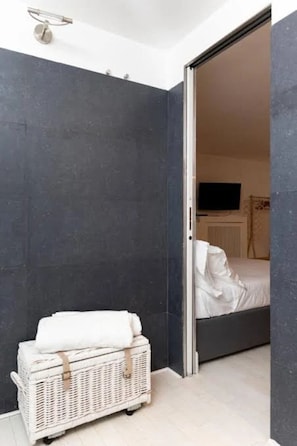 Ein modernes Badezimmer mit dunklen Wänden, einem weißen Korbwannenstuhl und einem sichtbaren Bett im angrenzenden Raum.